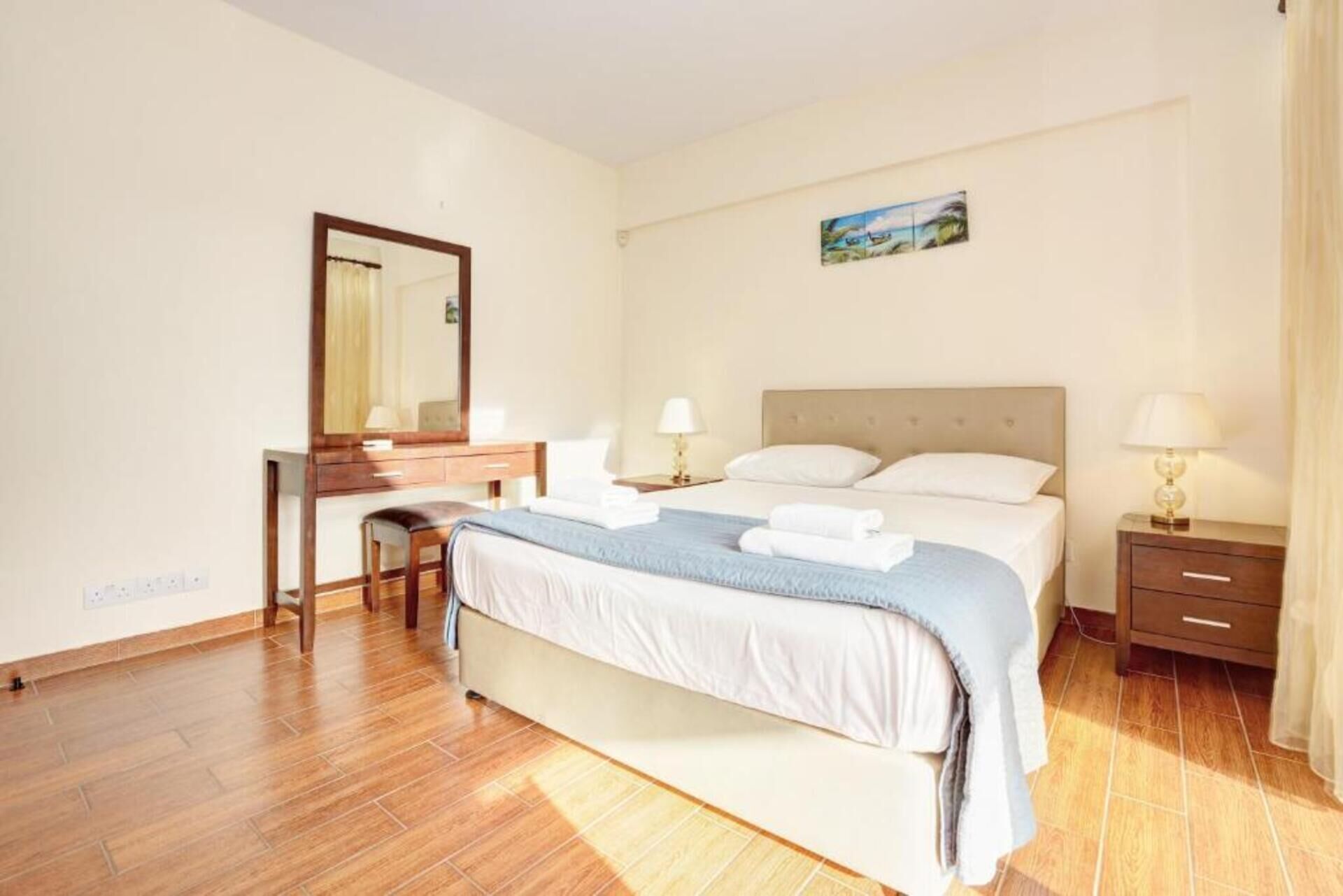 A bedroom with a bed, bedside tables, a desk, a chair, a mirror, and a painting on the wall.
