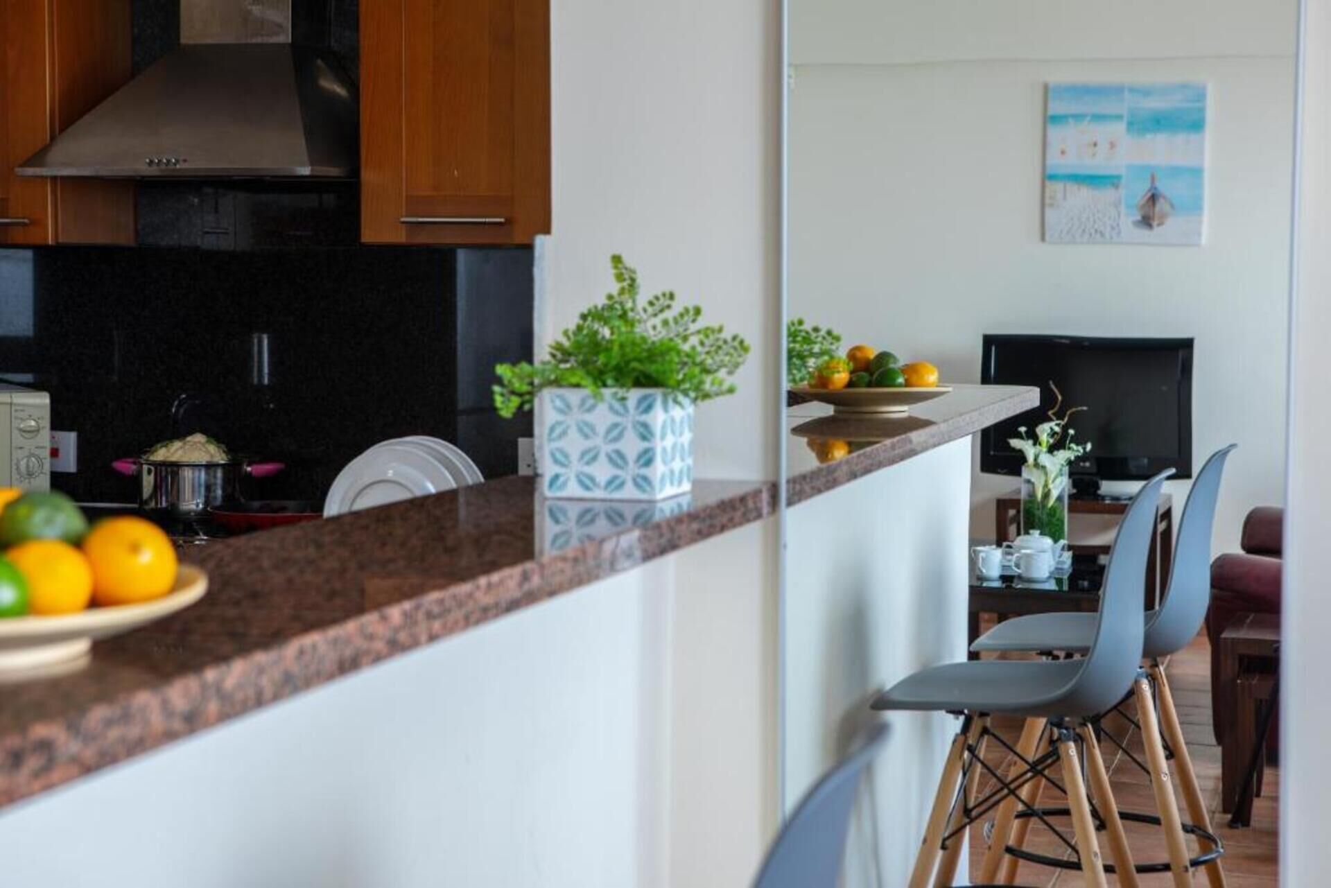 A modern kitchen with a granite countertop, a stainless steel range hood, and wooden cabinets. There is a potted plant on the counter and a dining area with a TV and chairs.