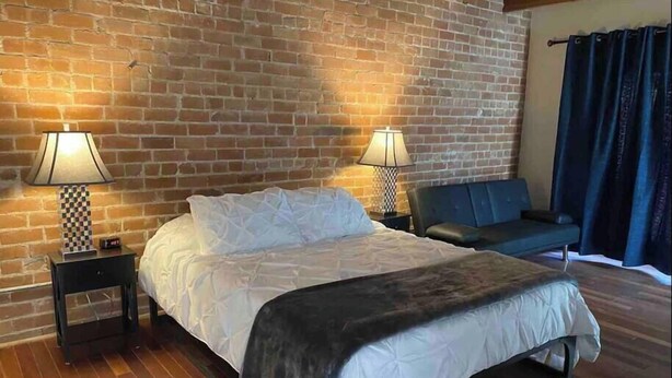 A bedroom with a brick wall, a bed with white bedding, two bedside tables with lamps, and a dark blue sofa. - Iron Horse Carraige Home & Parking (Tucson)