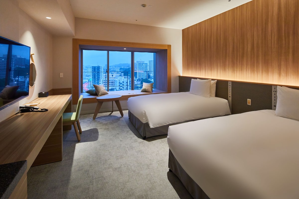 A hotel room with two beds, a desk, and a large window offering a city view.