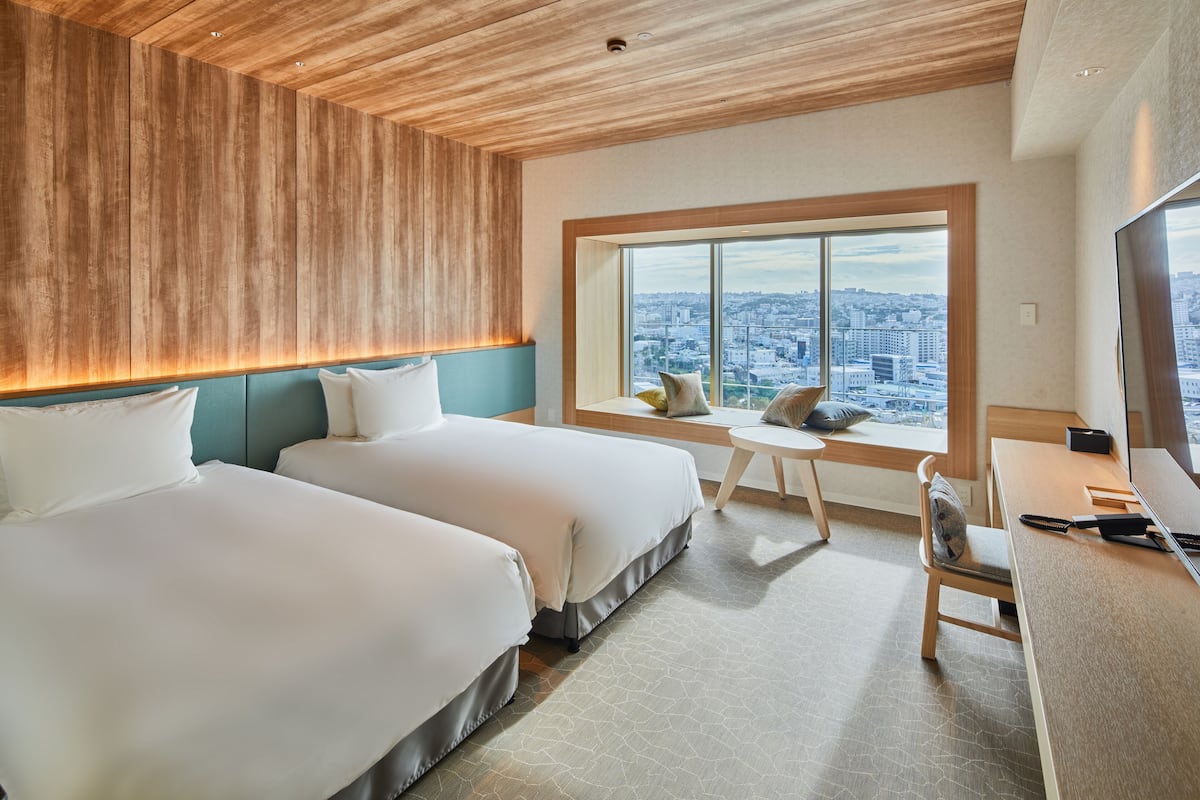 A hotel room with two beds, a desk, and a large window offering a city view.
