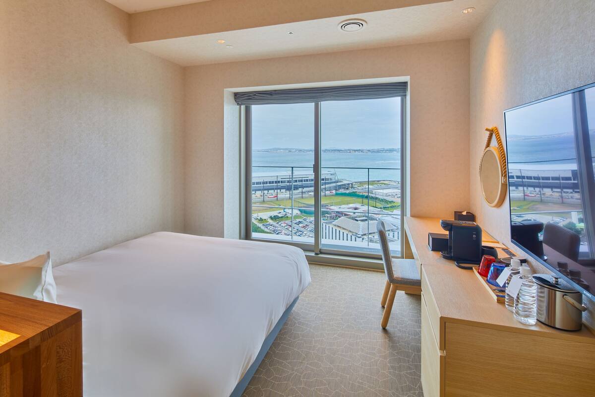 A hotel room with a bed, a desk with a computer, a chair, and a large window offering a view of the beach and sea.