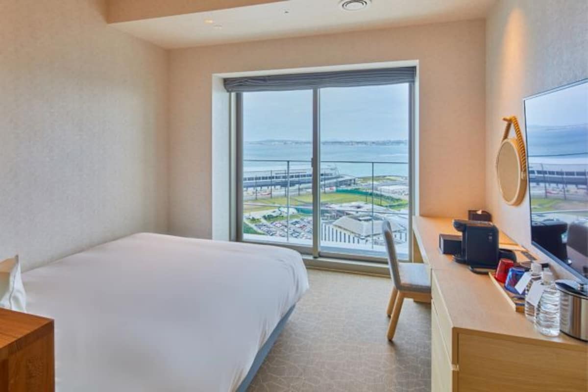 A hotel room with a bed, a desk with a computer, a chair, and a large window offering a view of a coastal area.
