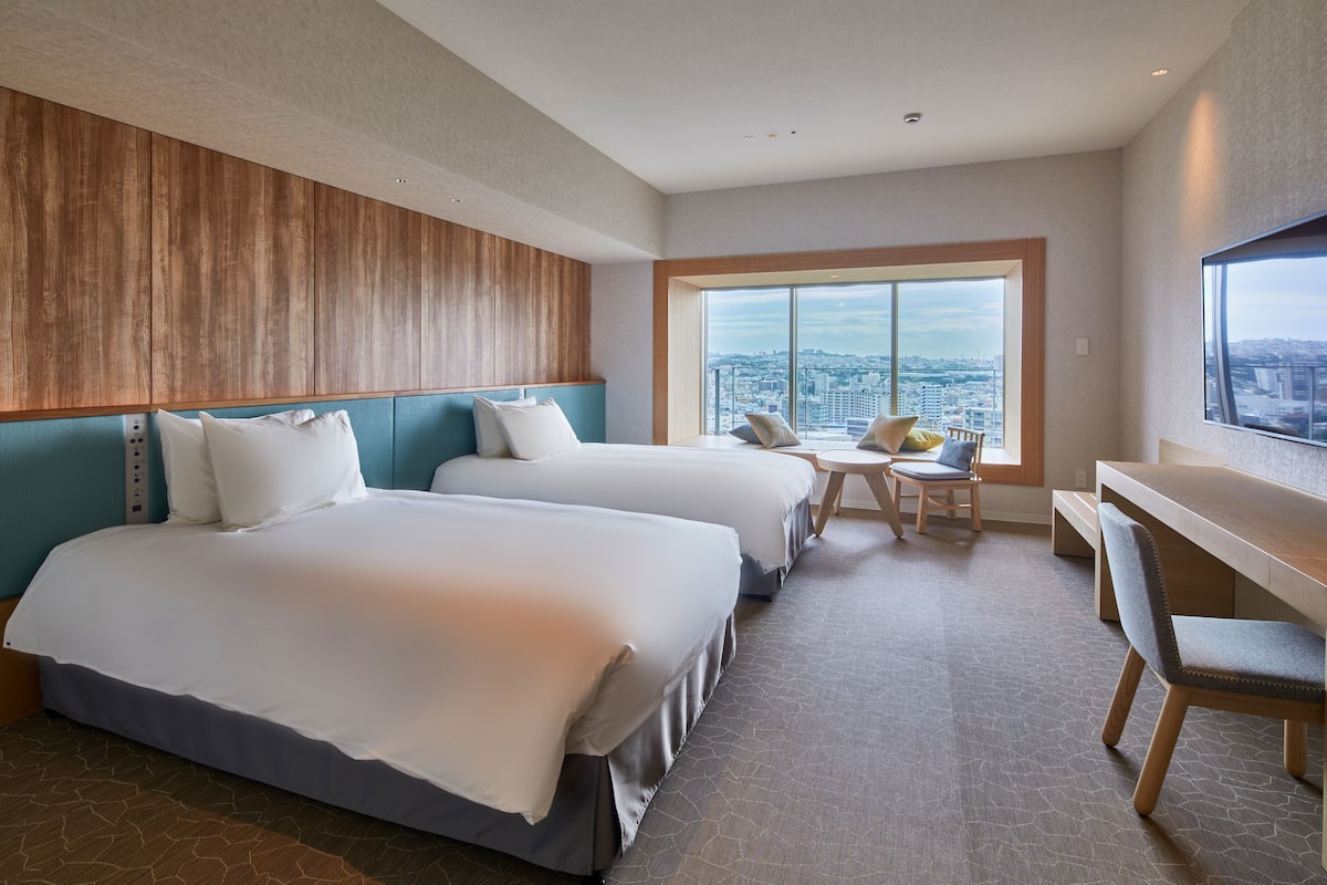 A hotel room with two beds, a desk, and a large window offering a city view.