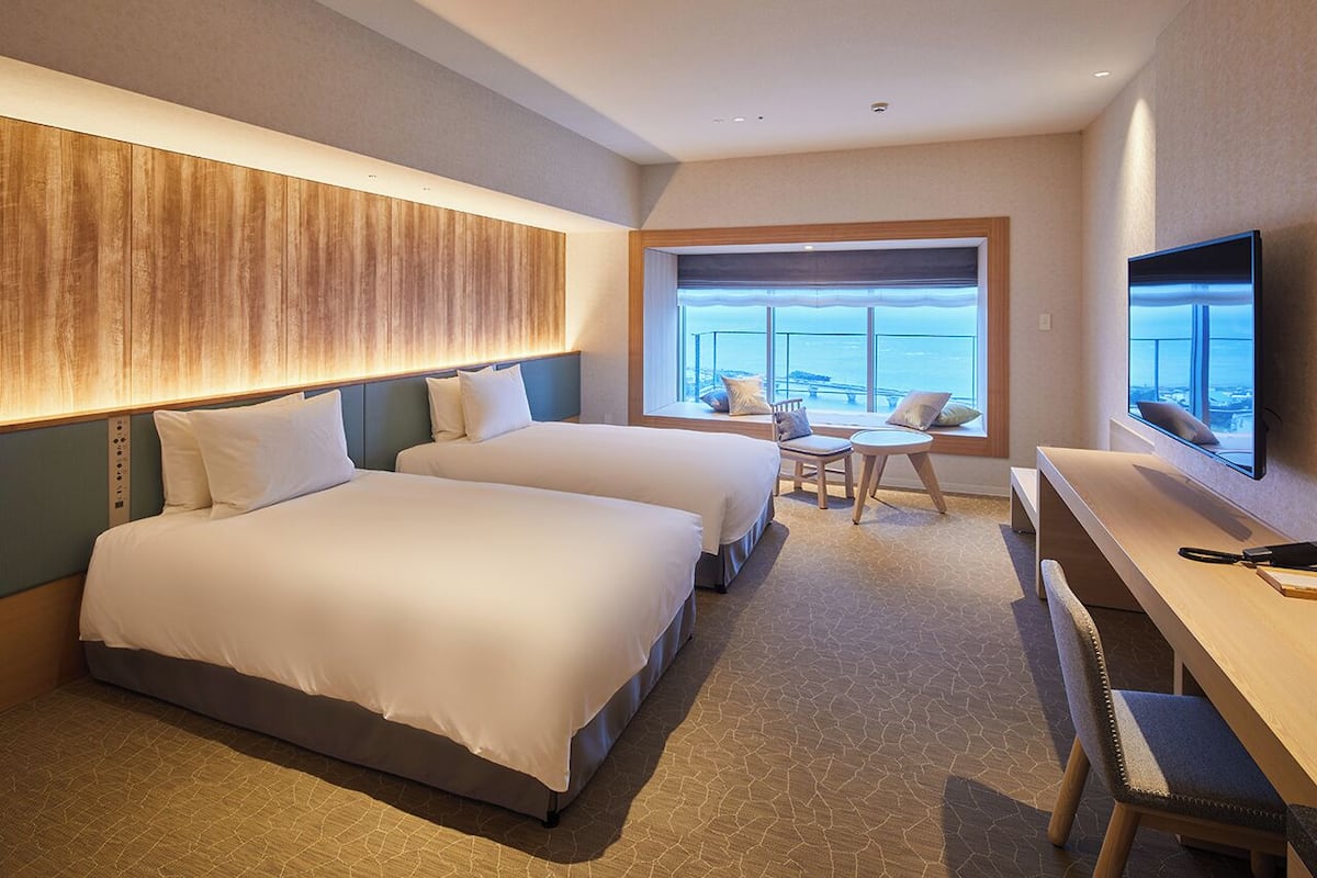 A hotel room with two beds, a desk, a TV, and a large window with a view.