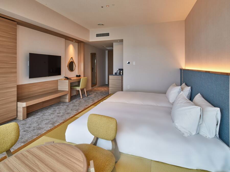 A hotel room with a large bed, a TV mounted on the wall, a desk with a chair, and a small seating area with a table and chairs.