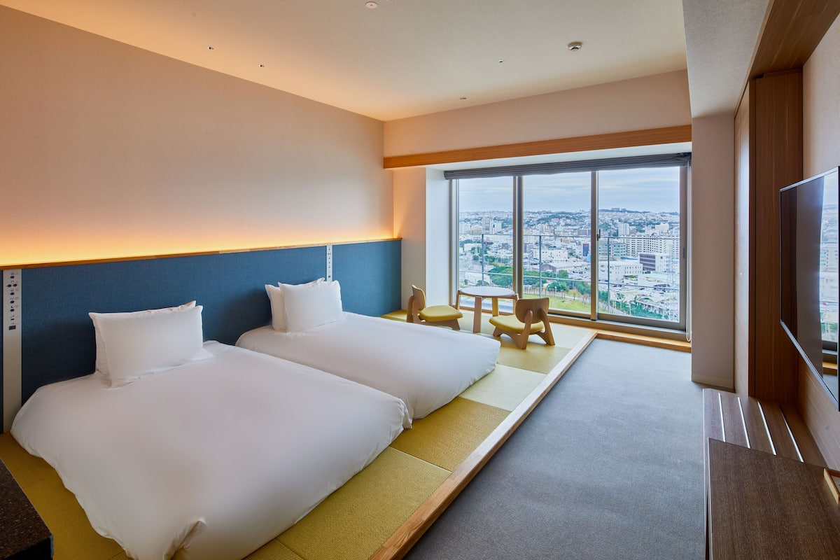 A hotel room with two beds, a TV, and a large window offering a city view.