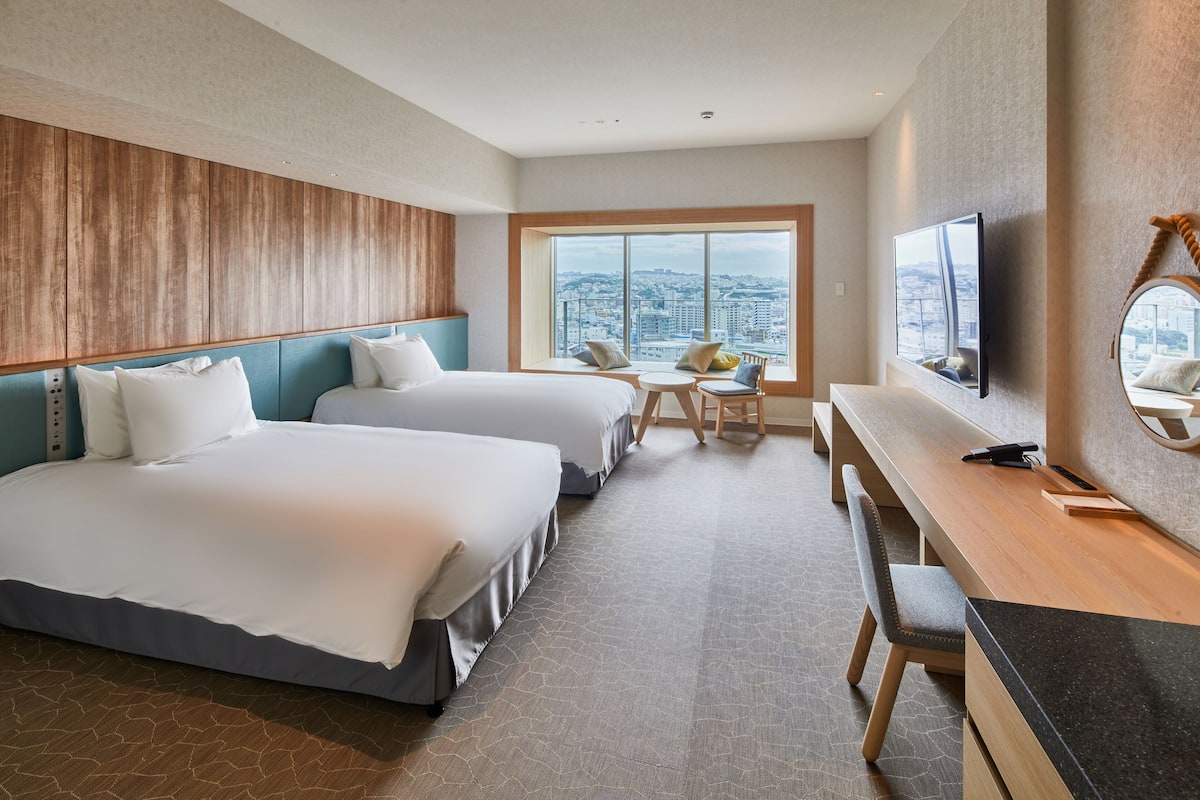 A hotel room with two beds, a desk, a chair, a TV, and a large window with a city view.