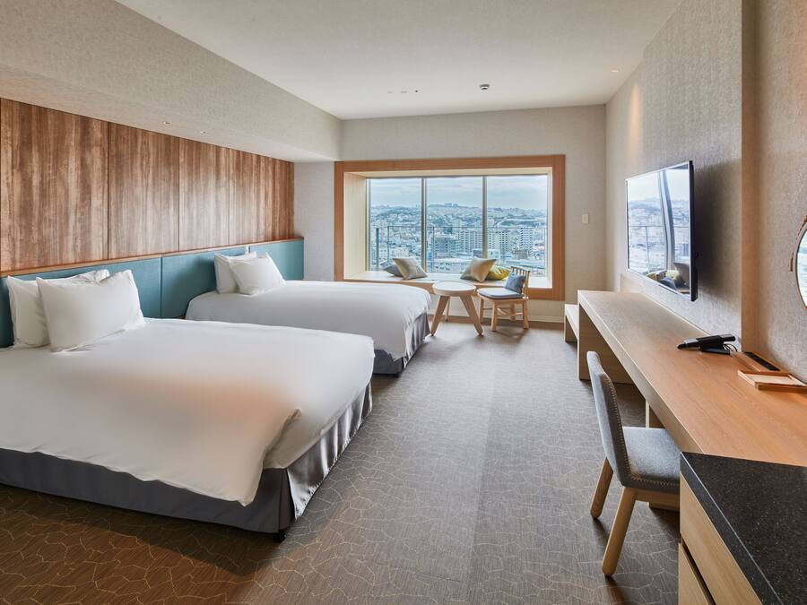A hotel room with two beds, a desk, a chair, a TV, and a large window with a city view.