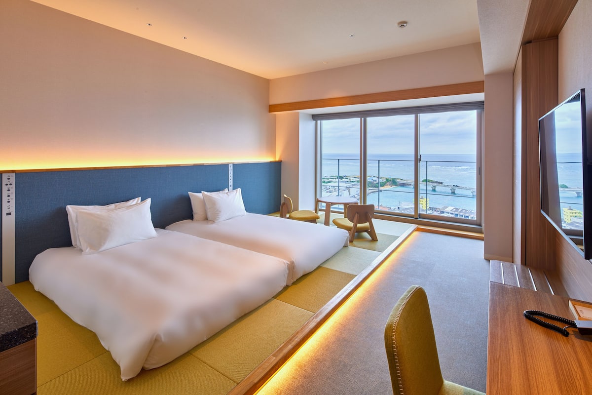 A hotel room with two beds, a desk, a chair, and a large window offering a view of the sea.