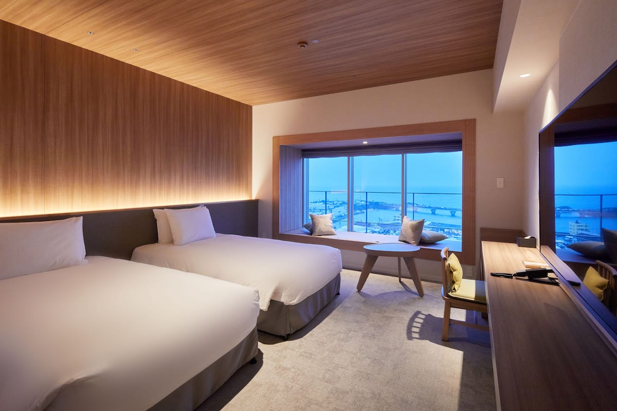 A modern hotel room with two beds, a small table, and a large window offering a view of the city.