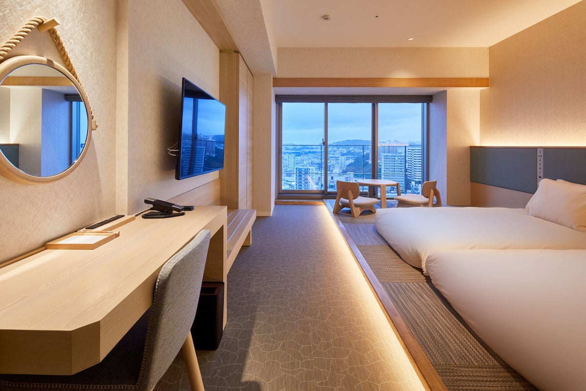 A hotel room with a desk, a bed, a TV, and a large window with a city view.