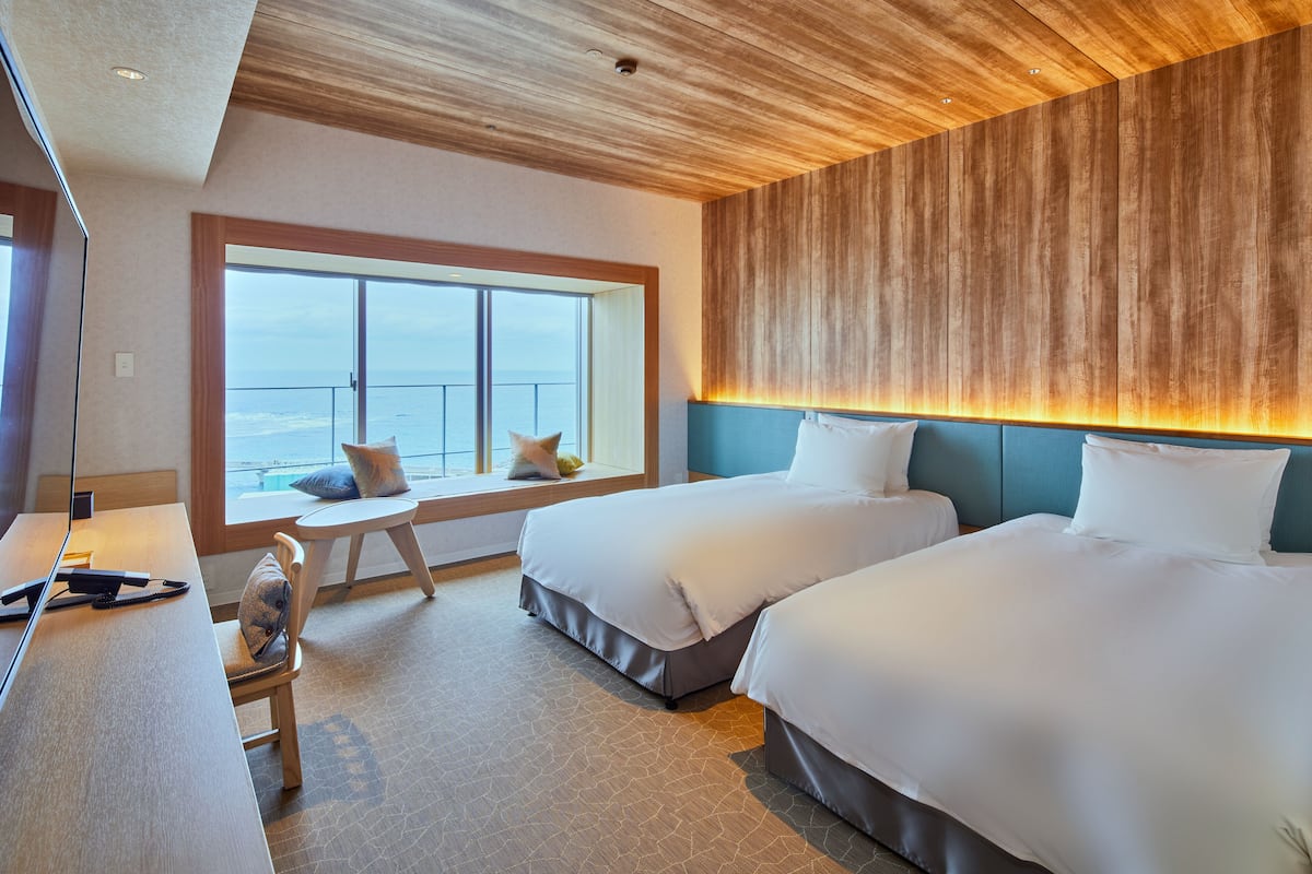 A hotel room with two beds, a desk, a chair, and a large window with a view of the ocean.