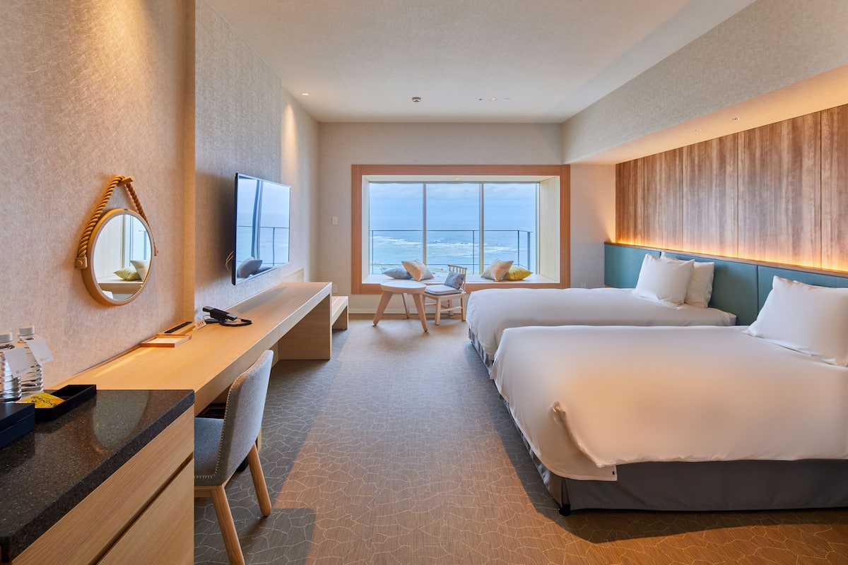 A hotel room with a desk, two beds, a TV, and a large window with a view of the ocean.