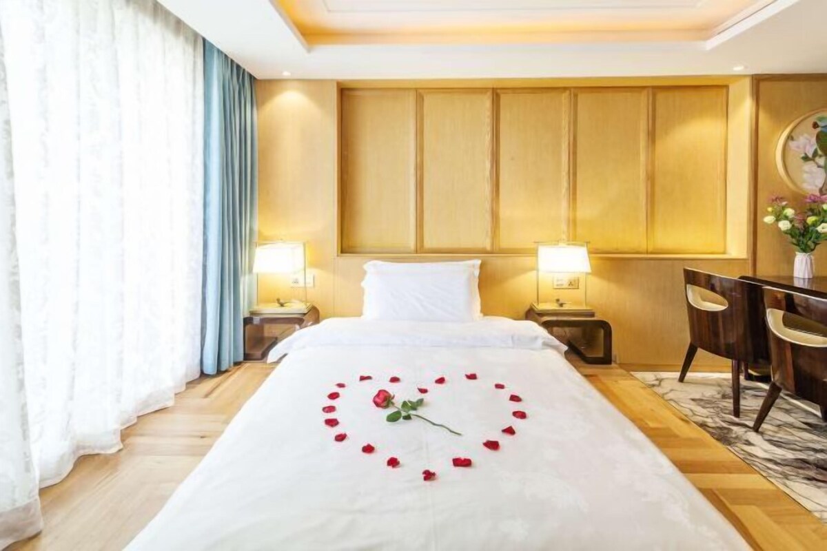 A hotel room with a bed, two bedside lamps, a wooden headboard, a desk with chairs, and a vase with flowers.