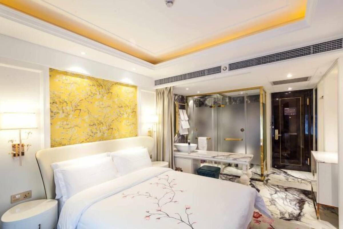 A hotel room with a bed, a vanity with a mirror, a bathroom, and a yellow wall with a floral pattern.
