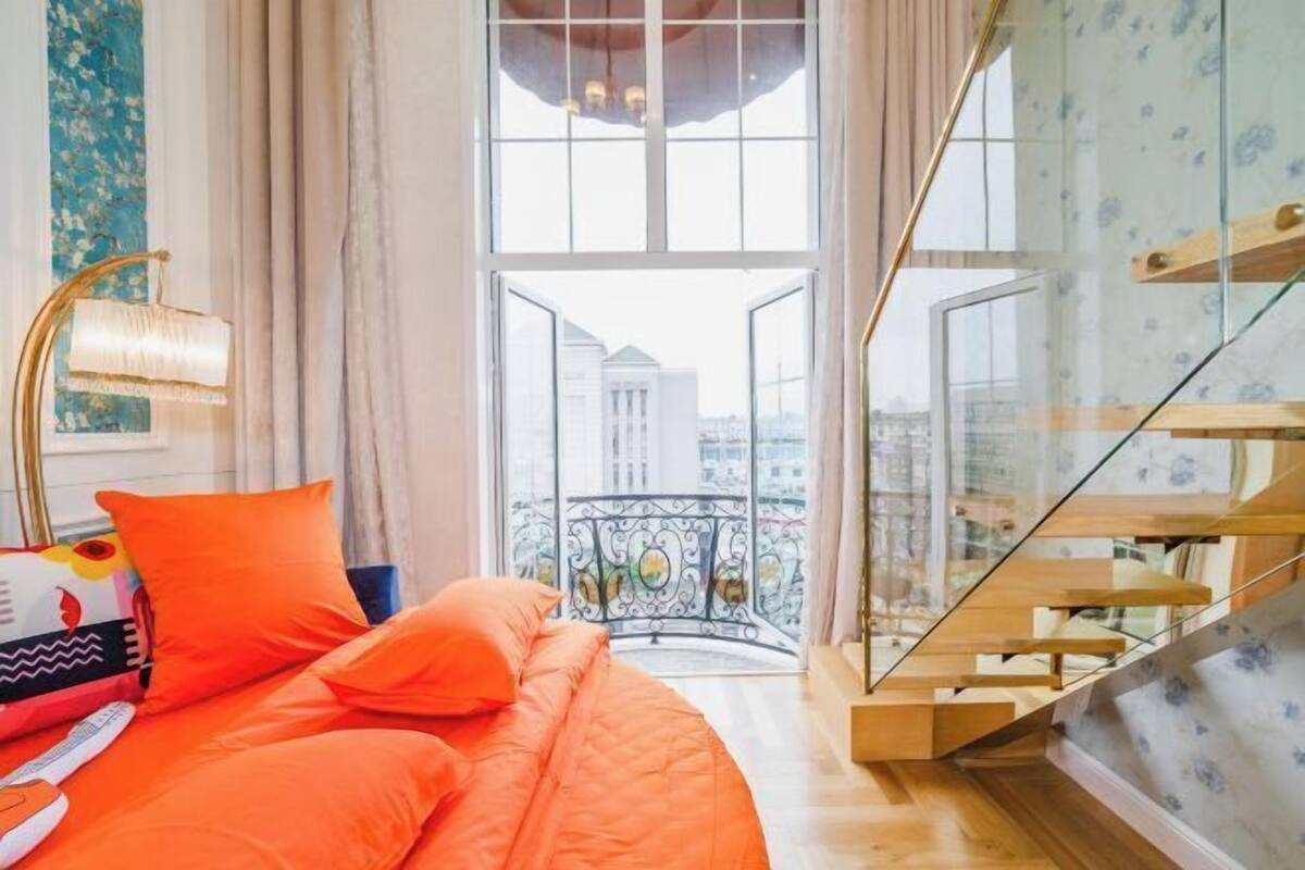 A bedroom with a bed, a window with a view of buildings, a staircase, and a balcony with a railing.