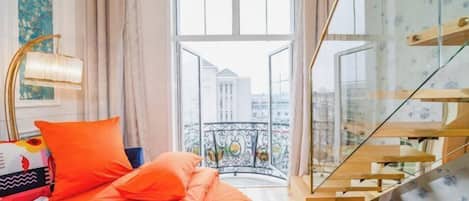 A bedroom with a bed, a window with a view of buildings, a staircase, and a balcony with a railing.