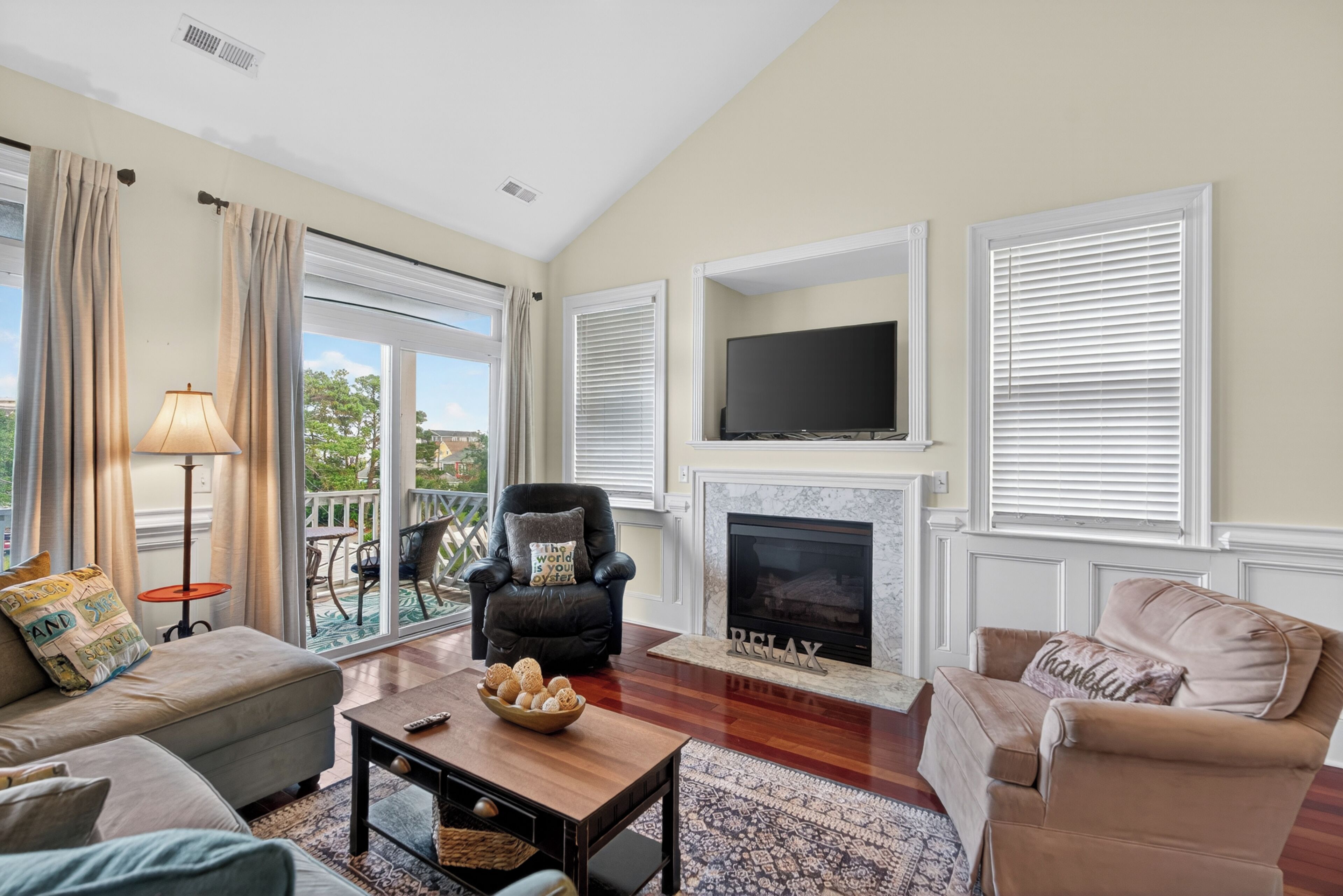 A living room with a sofa, armchair, fireplace, television, and a balcony with outdoor furniture.