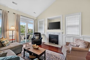 A living room with a sofa, armchair, fireplace, television, and a balcony with outdoor furniture.