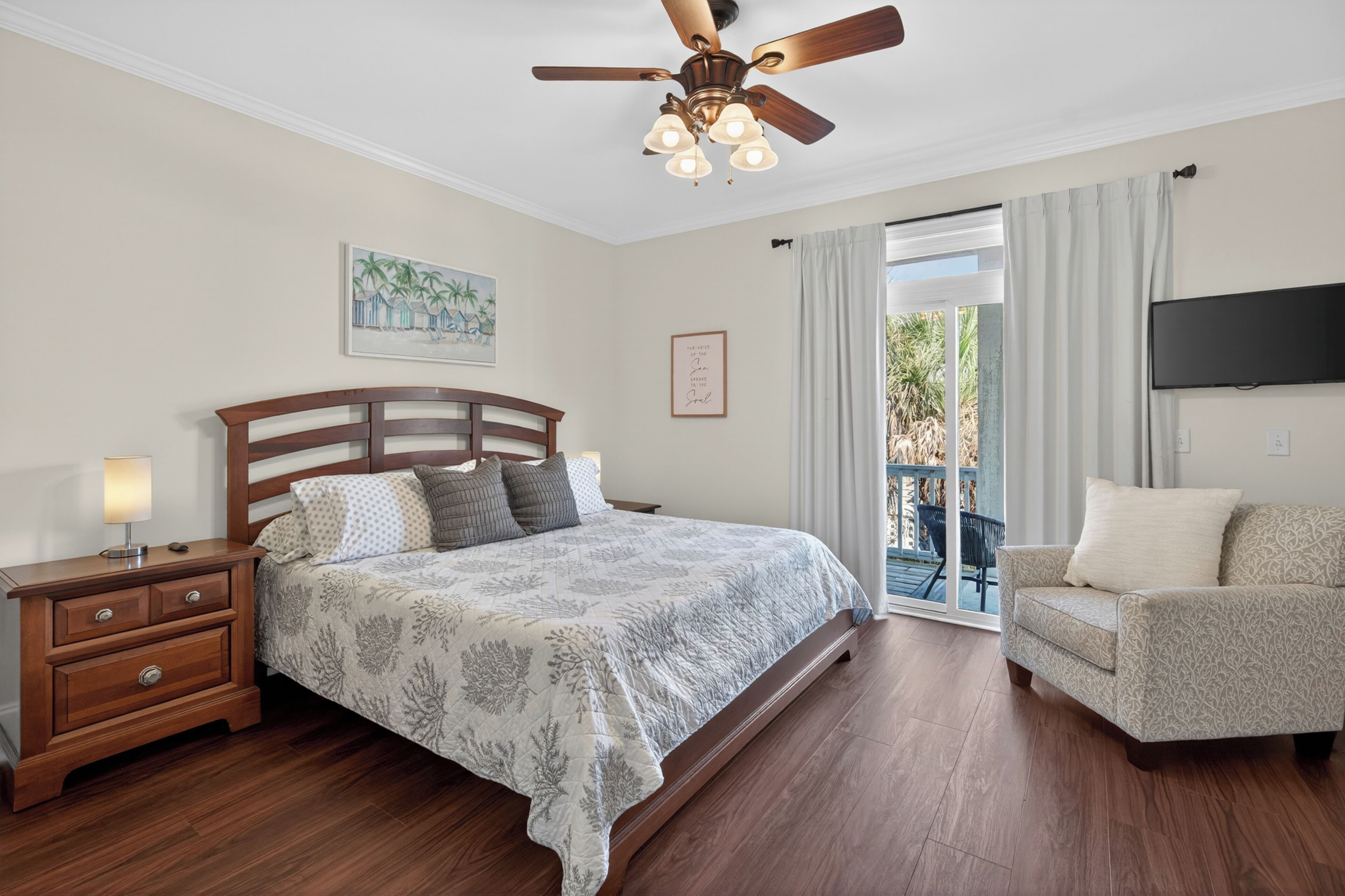 A bedroom with a bed, wooden nightstand, ceiling fan, artwork, and a TV on the wall.