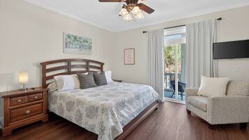 A bedroom with a bed, wooden nightstand, ceiling fan, artwork, and a TV on the wall.