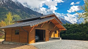 Une cabane en bois avec un toit en pente, une voiture garée à l’intérieur, et en arrière-plan, une vue sur des montagnes enneigées.