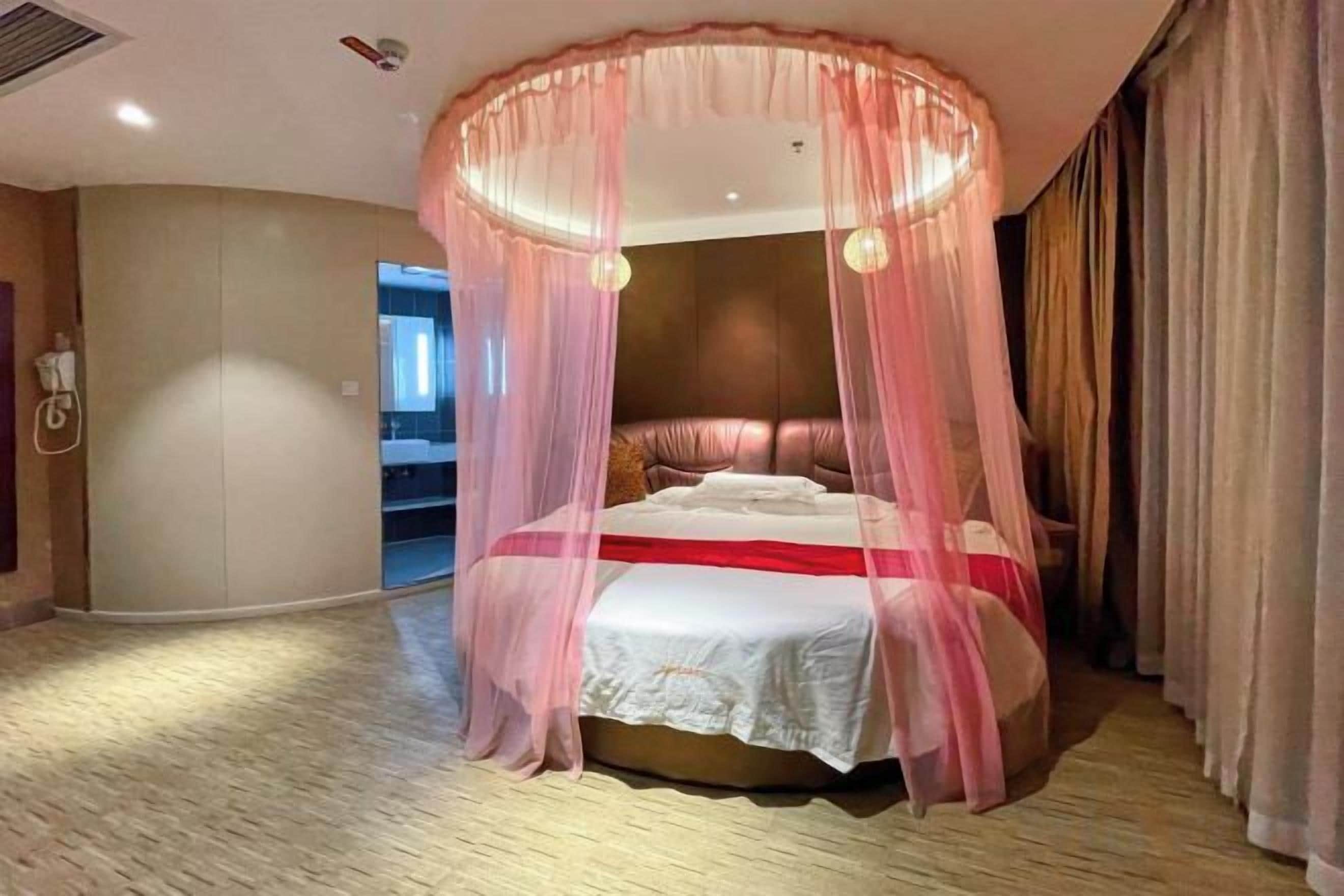 A four-poster canopy bed with a white bedspread and red accents, a wooden headboard, and a view of a hallway.