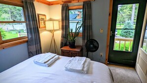 A bedroom with a bed, two towels, a standing lamp, a potted plant, a fan, and a view of a balcony with greenery. - Your Sunny Bungalow Awaits! (Big Indian)
