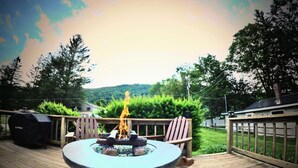 A deck with a fire pit, a grill, and a view of a forested hill. - Your Sunny Bungalow Awaits! (Big Indian)