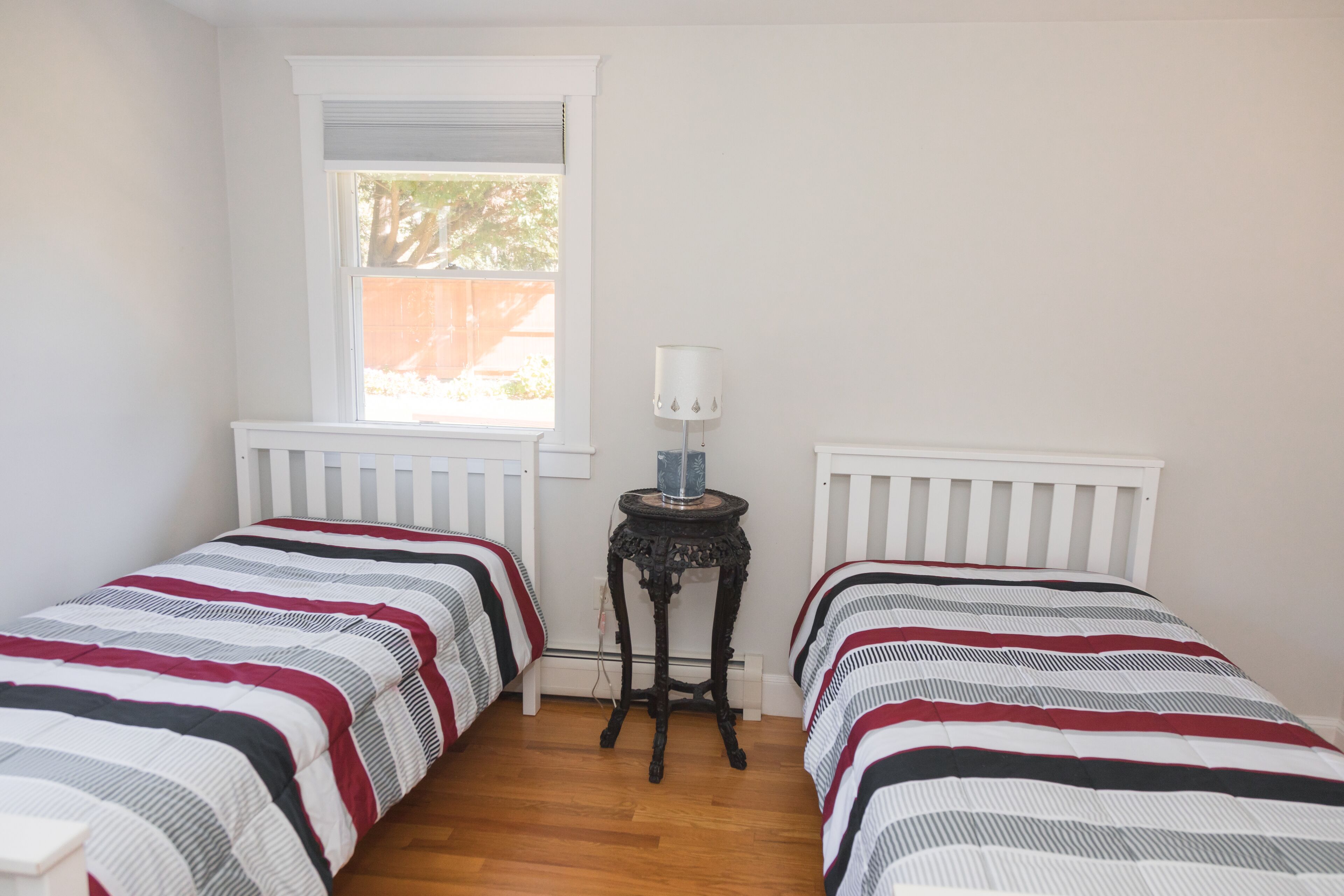 A bedroom with two single beds, a small table with a lamp, and a window with blinds.