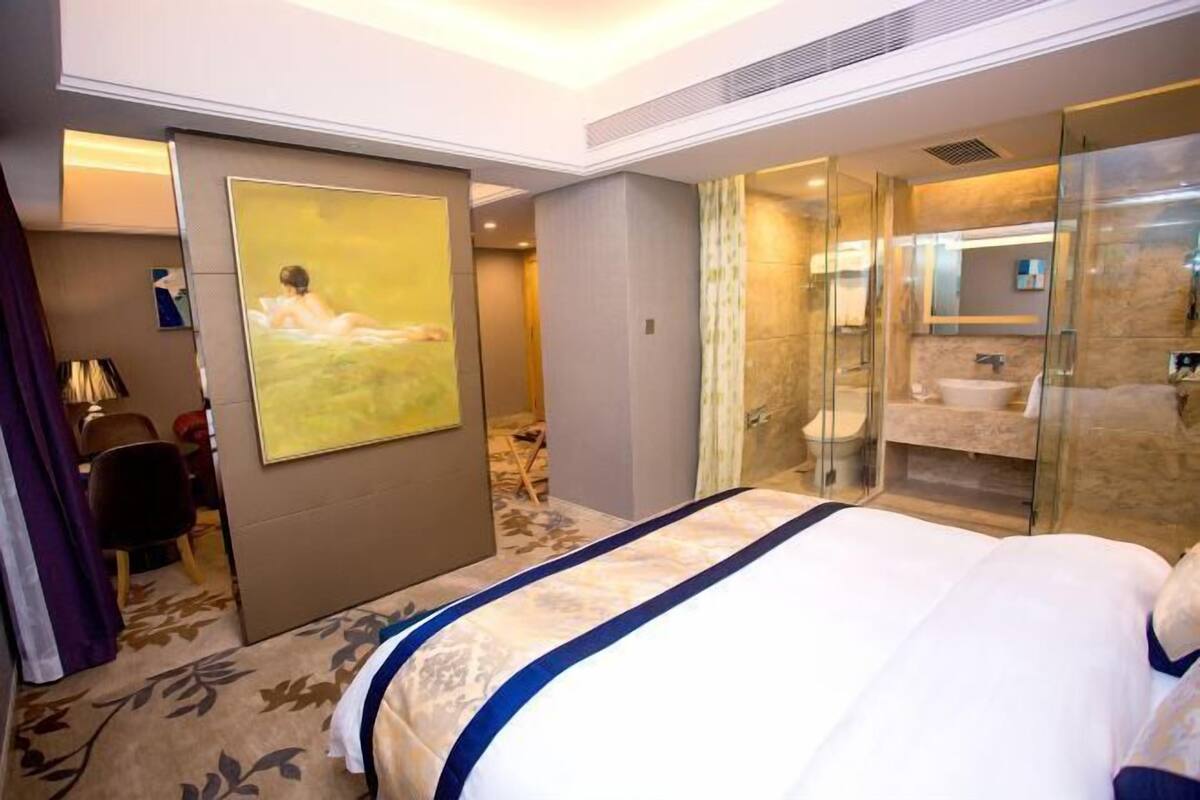 A hotel room with a large bed, a glass-enclosed shower, a painting on the wall, and a desk with a chair.