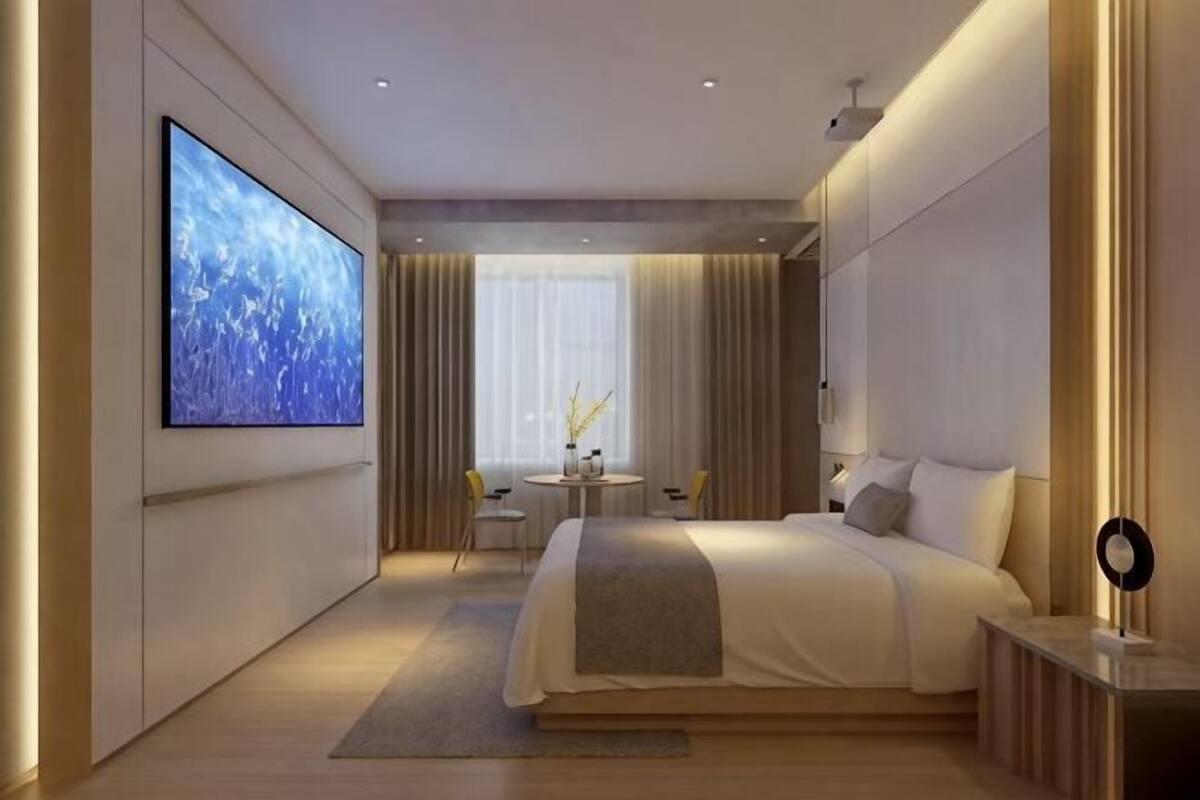 A modern hotel room with a large bed, a TV on the wall, a small table with chairs, and a window with curtains.