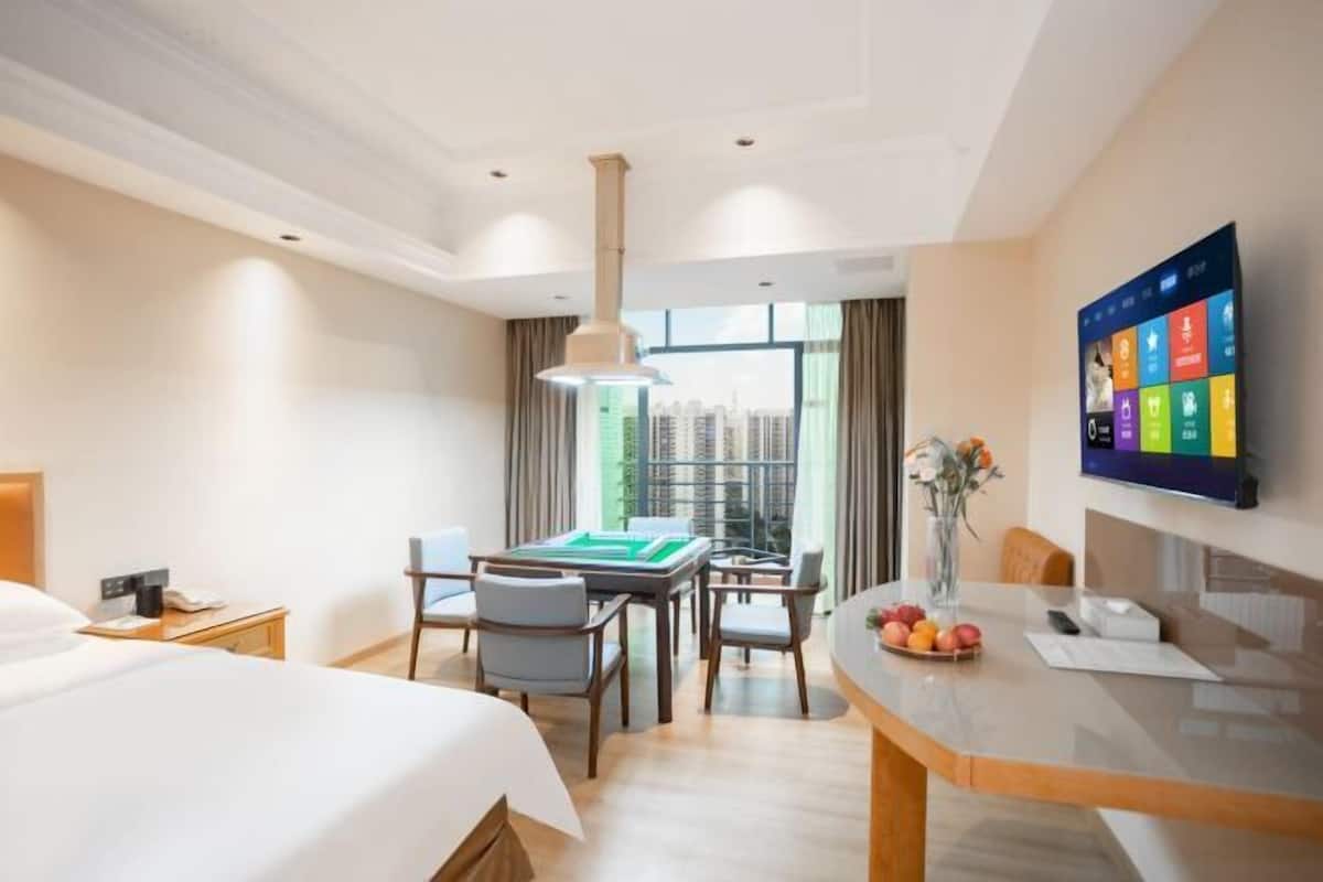 A hotel room with a bed, a desk with a computer, a dining table with chairs, a view of buildings through the window, and a TV mounted on the wall.
