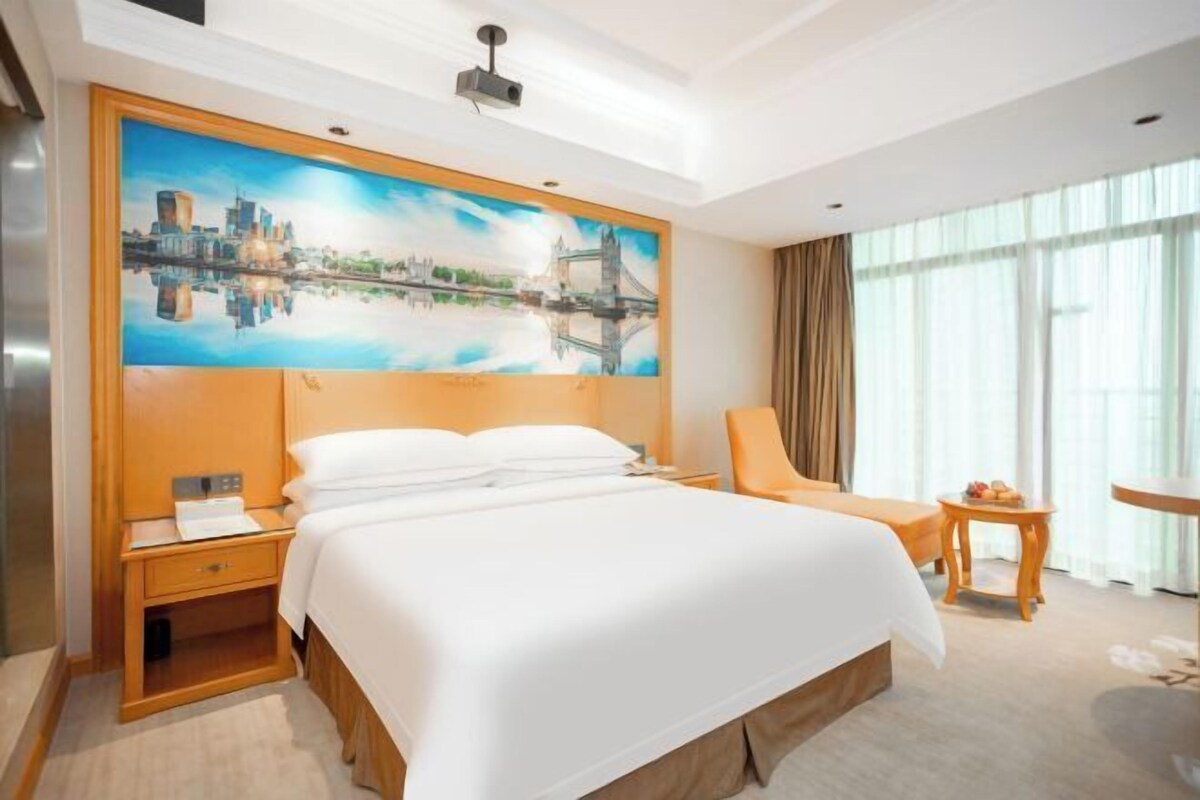 Ein Hotelzimmer mit einem großen Bett, einem Nachttisch, einem Sessel, einem kleinen Tisch und einem Gemälde einer Brücke.