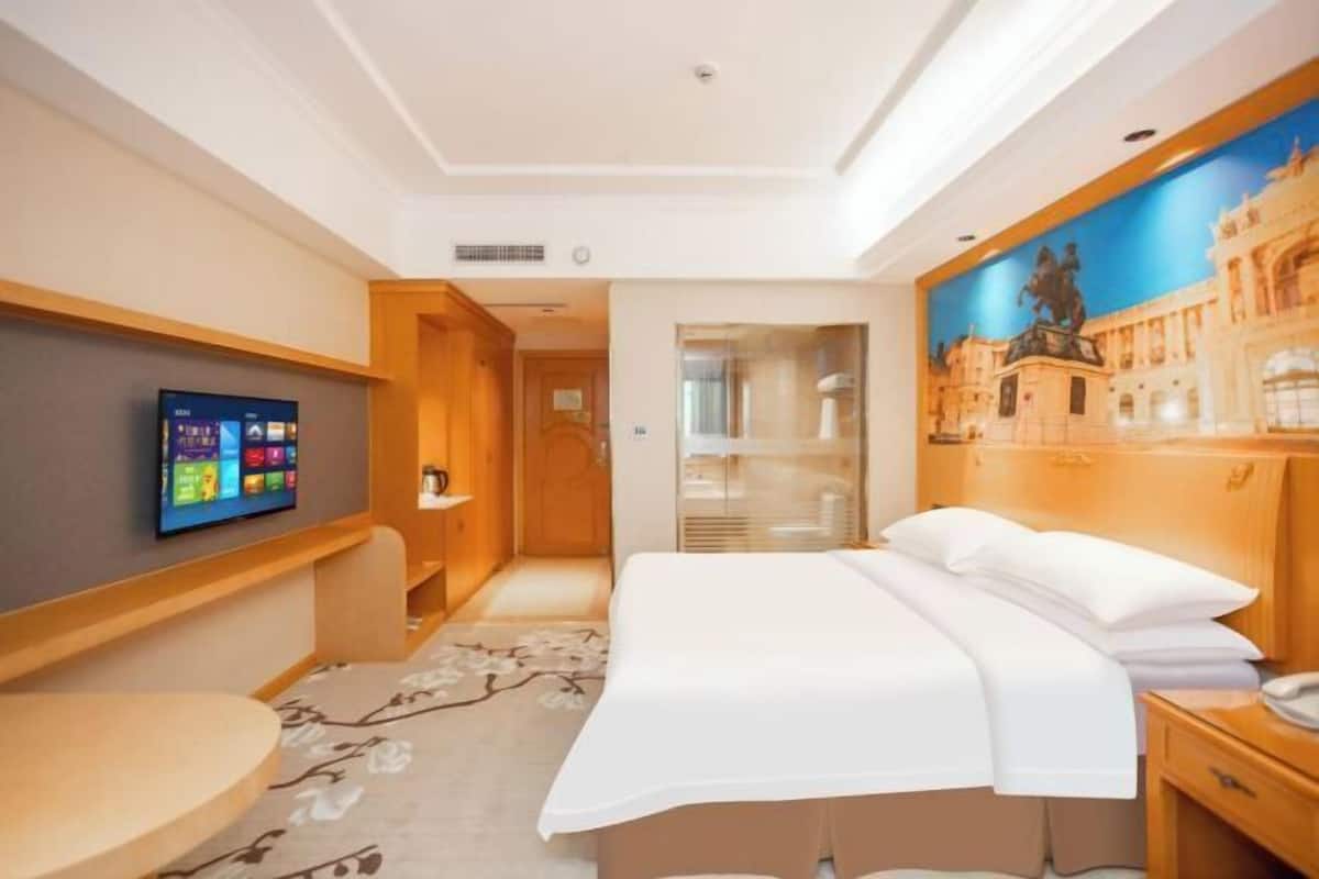 A hotel room with a bed, a TV mounted on the wall, a wooden headboard, and a painting of a cityscape.