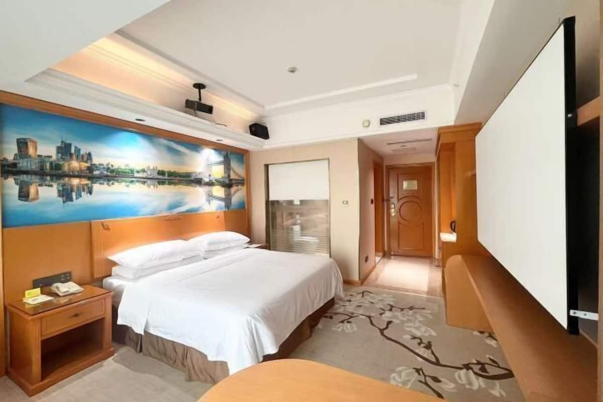 A hotel room with a bed, a nightstand, a painting of a cityscape, a television, and a door leading to another room.