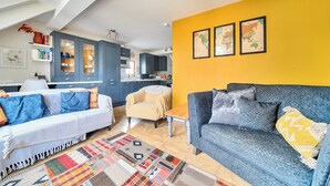 Un salon moderne avec un canapé, un fauteuil et une table basse. On aperçoit une cuisine à l’arrière-plan, avec des placards et un four à micro-ondes.
