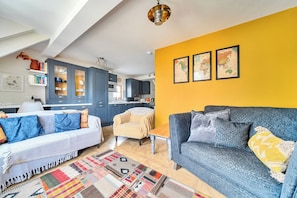 A living room with a sofa, armchair, and a coffee table. There is a kitchen in the background with cabinets and a dining area. - Newly Refurbished 2nd Floor Apartment Lil Nauti (Fowey)