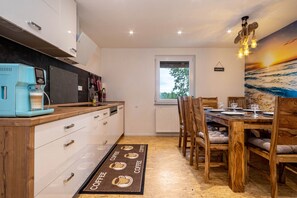 A modern kitchen with a coffee machine, wooden cabinets, a dining area with a wooden table and chairs, and a view of the outdoors through a window.