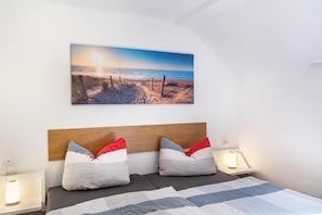 A bedroom with a bed, two bedside lamps, a headboard, and a beach scene painting on the wall.
