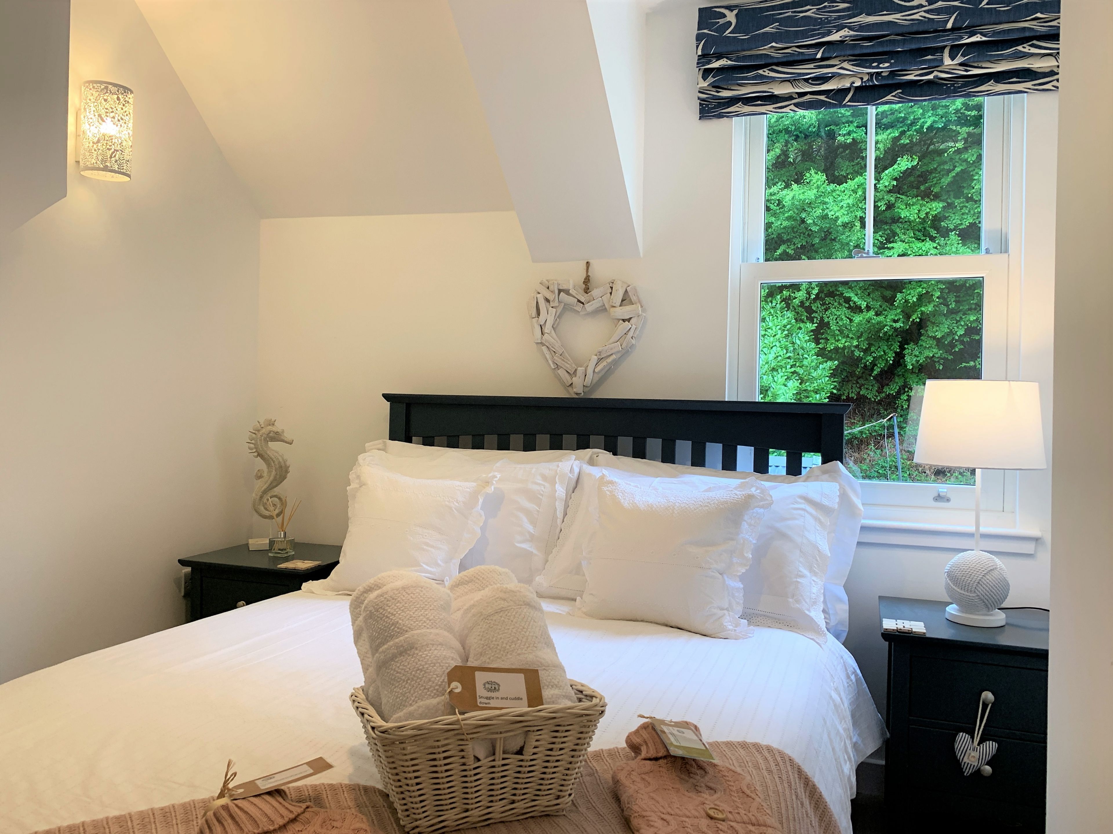 A neatly made bed with white linens and a basket of towels. There is a decorative wall piece above the bed and a window with a view of greenery outside.