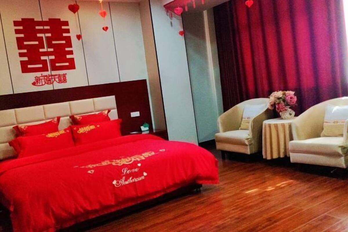 A hotel room with a red bedspread, a large bed, two armchairs, a small table, and a decorative flower arrangement.