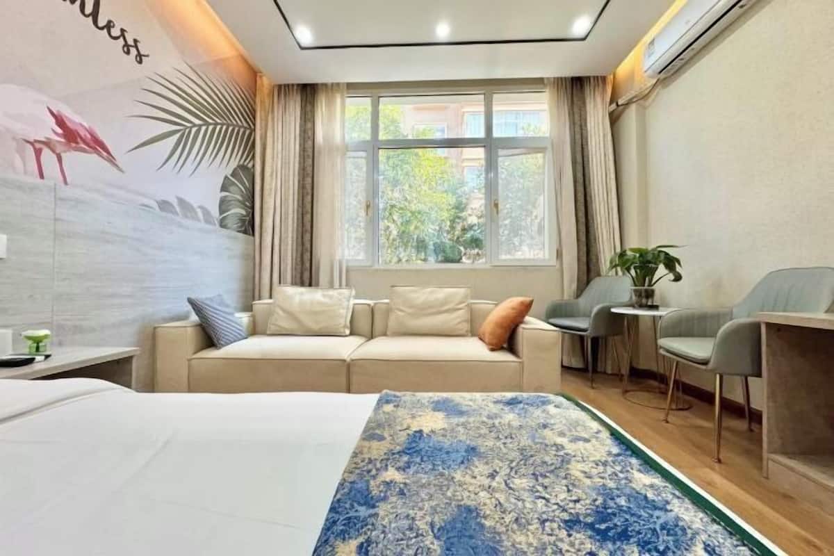 Ein modernes Hotelzimmer mit einem Bett, einem Sofa, einem kleinen Tisch und einem Sessel. Ein Fenster mit Vorhängen bietet Blick auf Grünflächen.