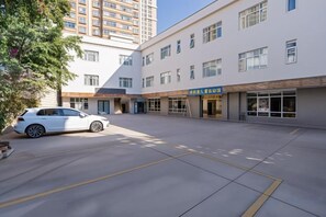A parking area with a white car, a building with multiple windows and a sign, and a tree. - Noah Hotel (Chengjiang)