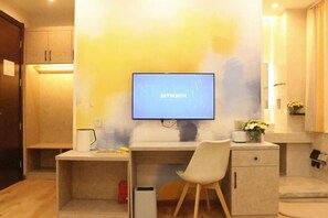 Una habitación moderna con televisión de pantalla plana en la pared, un escritorio con una silla, un mueble pequeño y una mesita de noche con lámpara.