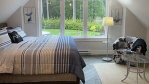 Een slaapkamer met een groot bed, een plafondventilator, een raam met uitzicht op groen en een tafeltje met een lamp.