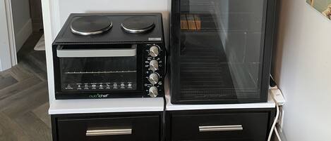 A compact kitchen with a microwave, oven, and a small stove. There is a shelf above the stove holding glasses and a teapot.