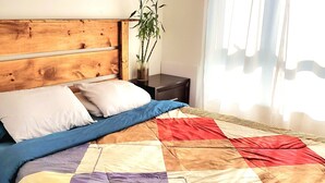 Quarto com cabeceira de madeira, cama com colcha colorida, mesa de cabeceira com uma planta e cortinas leves.
