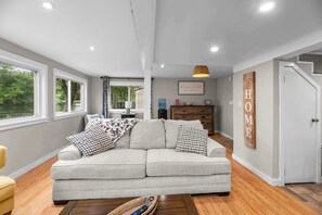 A spacious living room with a grey sofa, wooden flooring, and a view of a lake through large windows. - Little Landing Cottage | Lakefront · Dock · Kayaks (Winthrop)